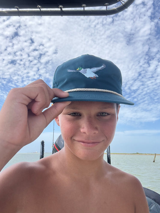 Person wearing a flying mallard duck vintage rope cap with a scenic design, standing on a boat with water and sky in the background.