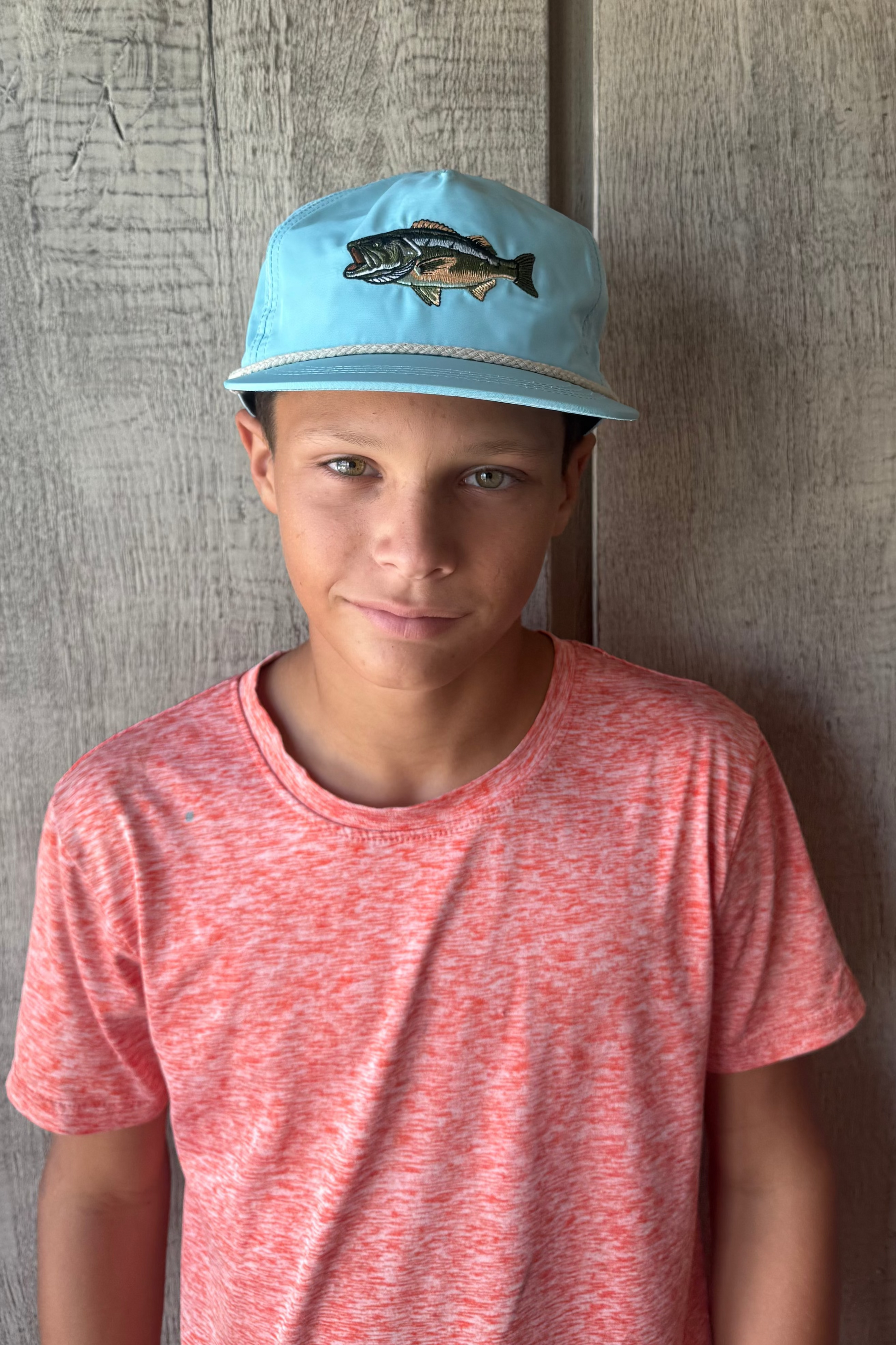 Teen boy wearing a blue cap with a fish design against a wooden wall.