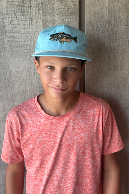 Teen boy wearing a blue cap with a fish design against a wooden wall.