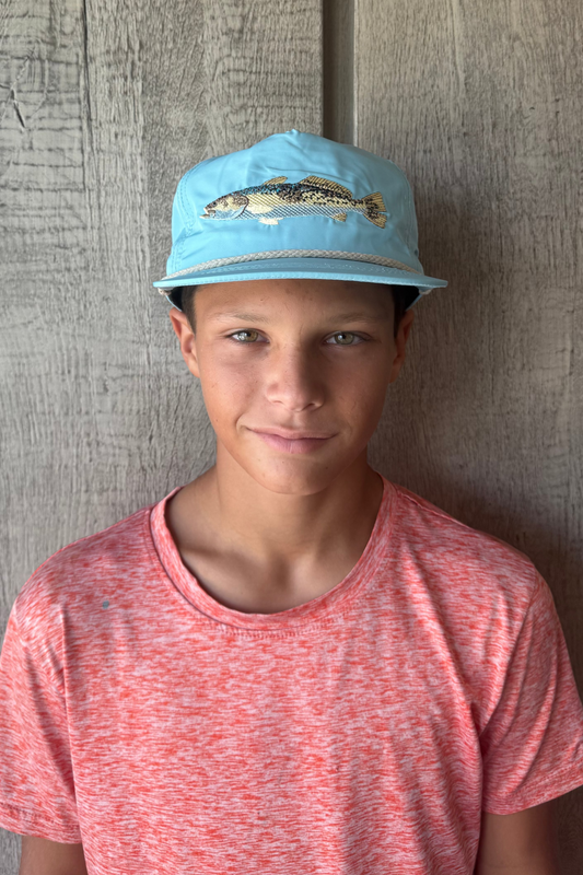 Teen boy wearing a blue cap with a speckled trout fish design against a wooden wall.