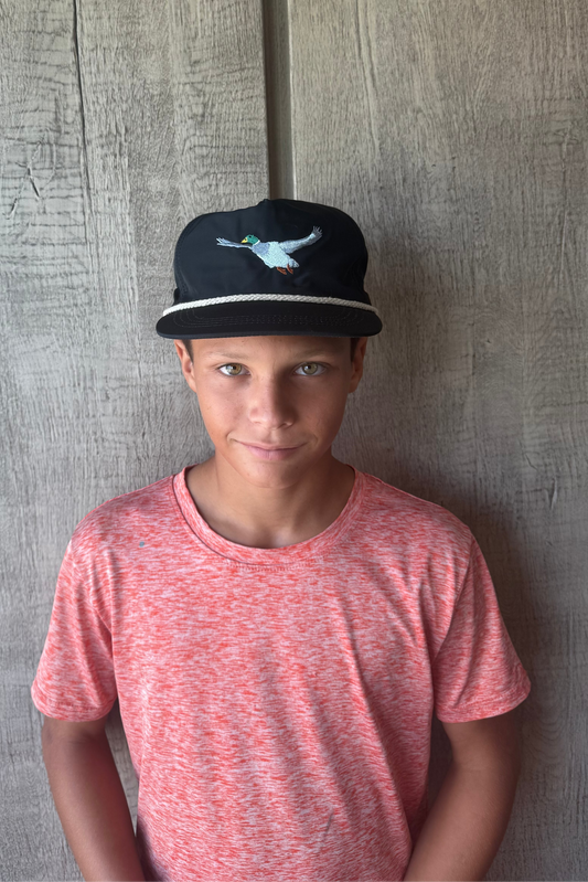Young boy wearing a black cap with a flying mallard duck outdoors and hunting rope cap against a textured gray oak wall
