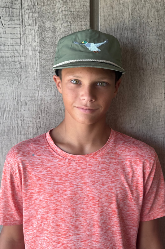 Teen boy wearing a red shirt and green rope cap with flying mallard duck embroidered on it against a textured gray wall
